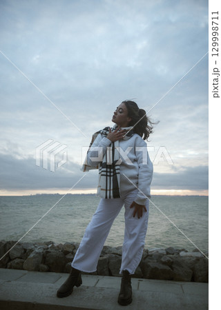 Beautiful young girl in a white tracksuit stands on the seashore in stormy weather Beautiful young girl in a white tracksuit stands on the seashore in stormy weather 129998711