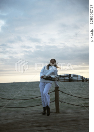 Beautiful young girl in a white tracksuit stands on the seashore in stormy weather Beautiful young girl in a white tracksuit stands on the seashore in stormy weather 129998717