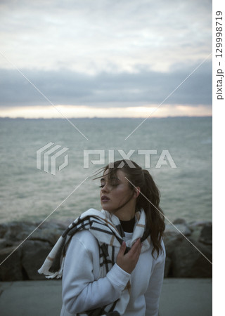 Beautiful young girl in a white tracksuit stands on the seashore in stormy weather Beautiful young girl in a white tracksuit stands on the seashore in stormy weather 129998719