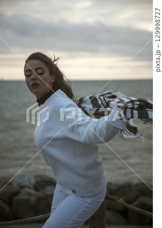Beautiful young girl in a white tracksuit stands on the seashore in stormy weather 129998727
