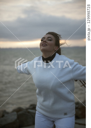 Beautiful young girl in a white tracksuit stands on the seashore in stormy weather Beautiful young girl in a white tracksuit stands on the seashore in stormy weather 129998728