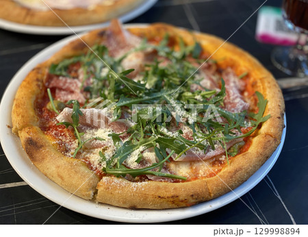 A mouth-watering Italian pizza with prosciutto, arugula, and parmesan cheese on a white plate A mouth-watering Italian pizza with prosciutto, arugula, and parmesan cheese on a white plate 129998894