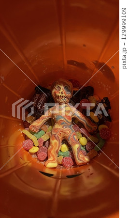 A vibrant, marbled human-like figure, resembling a voodoo doll, rests amidst an assortment of colorful Halloween candies and chocolate bars inside an orange bucket, viewed from above with scattered. 129999109
