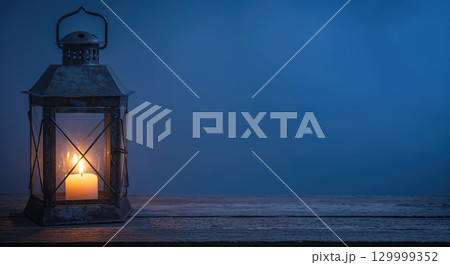 An old, rustic metal lantern with frosted glass panes and a lit candle inside sits on a wooden surface against a dark blue background, offering warm light and copy space. 129999352