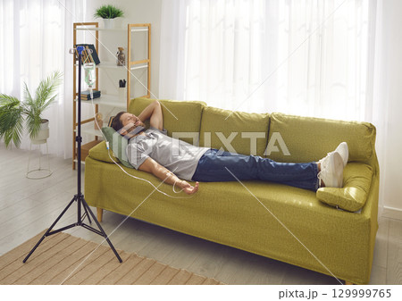 Smiling man lying on the couch at home while receiving IV drip infusion and vitamin therapy. 129999765