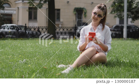 A cheerful young woman is enjoying her time relaxing on the grass while using her smartphone on a bright sunny day A cheerful young woman is enjoying her time relaxing on the grass while using her smartphone on a bright sunny day 130000187