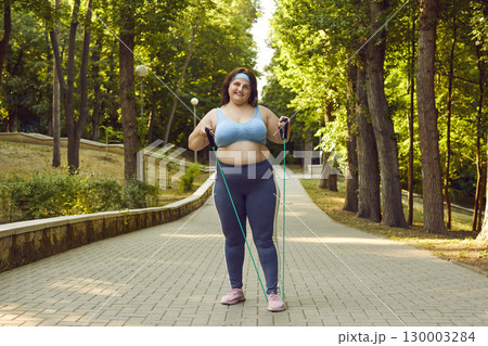 Fat woman doing sport exercises with rubber band standing in summer park outdoors. Fat woman doing sport exercises with rubber band standing in summer park outdoors. 130003284
