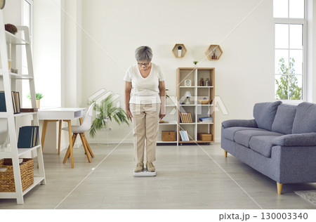 Senior woman standing on weight scales in the living room and looking down at figures 130003340