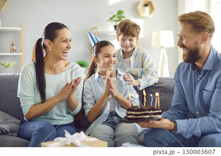 Happy girl in party hat celebrating birthday with family at home, looking at cake with candles Happy girl in party hat celebrating birthday with family at home, looking at cake with candles 130003607