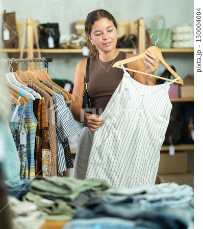Girl customer choose dress in store, and buyer carefully examines product. 130004004