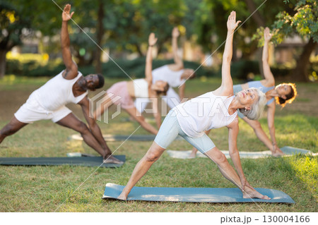 Multinational yoga enthusiasts with mature female coach perform Trikonasana in park Multinational yoga enthusiasts with mature female coach perform Trikonasana in park 130004166