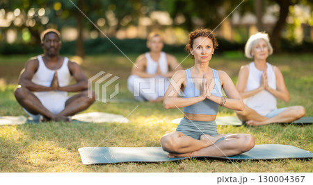 Multinational yoga enthusiasts with female coach perform padmasana in park 130004367