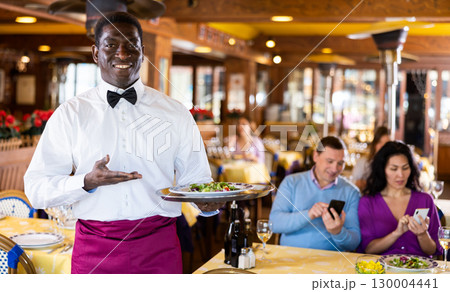 Positive African-american waiter with tray in restaurant 130004441