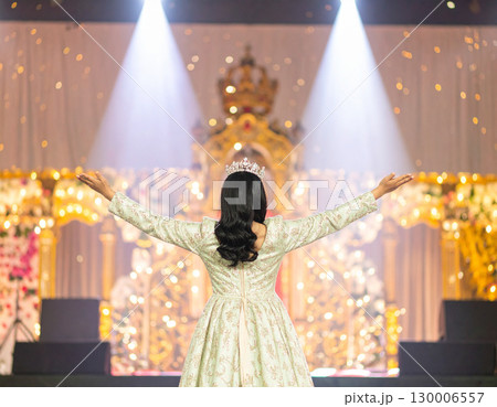 AI generated A radiant beauty queen stands at the frame edge gesturing toward a sparkling pageant stage with spotlights. The dazzling backdrop evokes elegance, competition and dream-come-true moments 130006557