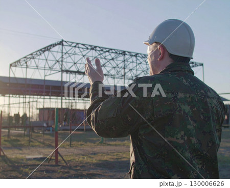 AI generated A uniformed soldier stands at the frame edge gesturing toward an active military training camp, showcasing discipline and teamwork. The background features obstacle courses and barracks 130006626
