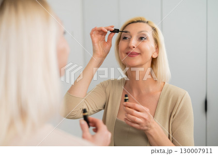 Elegant female using mascara wand to enhance lashes in front of mirror indoors 130007911