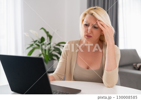 Middle aged blonde woman in beige clothes sadly looking at bill, sitting in cozy living room, concept of financial problems and household stress Middle aged blonde woman in beige clothes sadly looking at bill, sitting in cozy living room, concept of financial problems and household stress 130008585