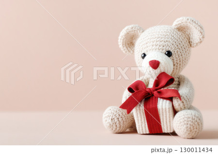 Adorable knitted teddy bear holding a small gift box with a red ribbon, set against a soft pastel background, perfect for celebrations and gifts 130011434