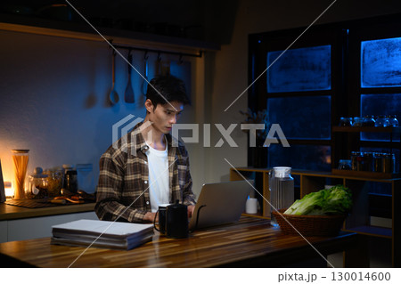 Concentrated freelancer sitting at kitchen counter, using laptop at midnight surrounded by documents 130014600