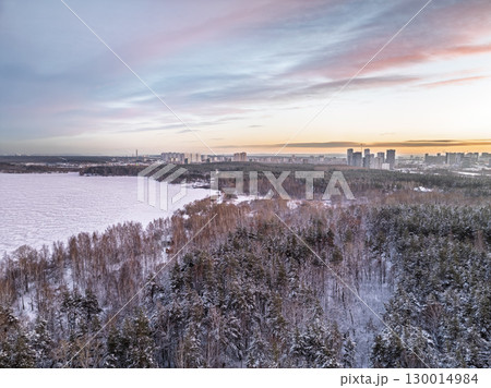 Snow-covered forest on lake shore with the first ice at sunset and the city on horizon, aerial view 130014984