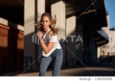 Young athletic woman exercising outdoors in sunlight near modern architecture 130016008