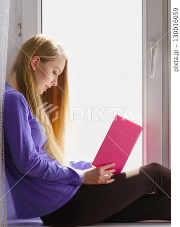 Woman sitting on window sill reading book at home 130016059