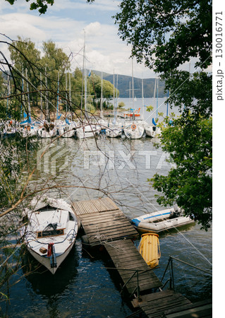 Sailboats are moored at wooden pier on calm lake, with lush green trees framing serene scene and rolling hills visible in distance under cloudy sky. Wooden pier hosting moored sailboats, cloudy sky Sailboats are moored at wooden pier on calm lake, with lush green trees framing serene scene and rolling hills visible in distance under cloudy sky. Wooden pier hosting moored sailboats, cloudy sky 130016771