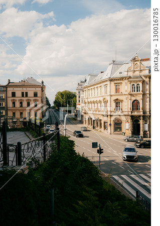 Cars driving along street, passing picturesque buildings under cloudy sky, with lush vegetation enhancing serene urban landscape. Street scene: vehicles moving past historic buildings 130016785