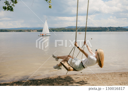 Blonde woman with sunglasses swinging on swing on beach enjoying summer by lake with sailboat in background, concept of freedom and summer vacation. Woman swinging near sailboat, relaxing by lake 130016817