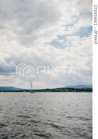 Sailboat gliding across lake, surrounded by cloudy sky and majestic mountains in background, creating serene and picturesque landscape filled with tranquility. Sailboat gliding across misty lake 130016822