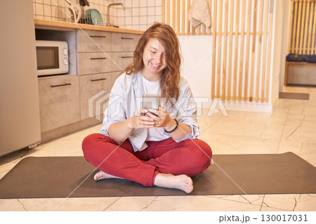 Young caucasian female in kitchen sitting on mat using smartphone, relaxed home 130017031