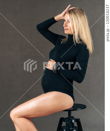 Pregnant woman in thoughtful pose sitting on stool, displaying serene expression while gently holding her belly. Wearing a black outfit, she expresses beauty and anticipation. 130018237