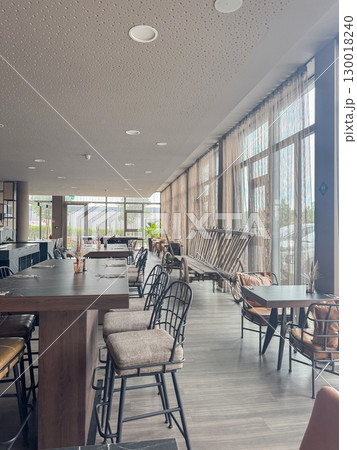 Modern cafe, hotel interior with empty tables, high stools, and large windows. Neutral tones and simple decor create a welcoming and calm ambiance, ideal for relaxation or work Modern cafe, hotel interior with empty tables, high stools, and large windows. Neutral tones and simple decor create a welcoming and calm ambiance, ideal for relaxation or work 130018240