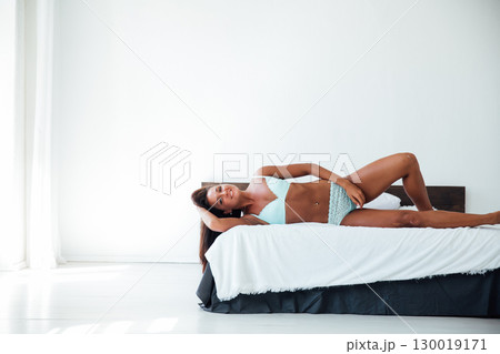 Tanned woman in light lingerie lying in bedroom on bed 130019171