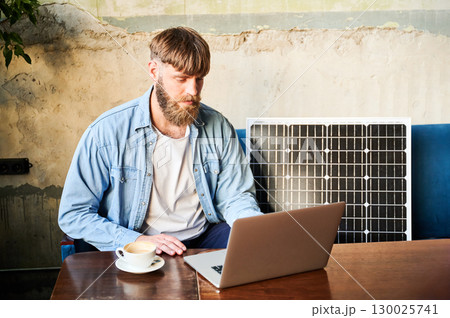 Bearded man works on laptop at wooden table, with cup of coffee. Solar panel nearby, emphasizing sustainable eco-friendly workspace that combines technology and sustainability in cozy environment. 130025741