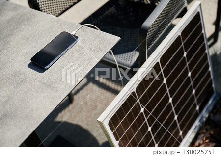 Smartphone connected to solar panel on outdoor table, emphasizing practical use of solar energy for charging devices. Integration of renewable energy, showcasing portable solar technology. 130025751
