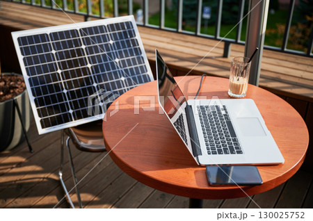 Laptop, smartphone and glass of iced coffee on round wooden table on sunny balcony. Solar panel mounted nearby, showcasing modern, eco-friendly workspace that combines technology and sustainability. 130025752