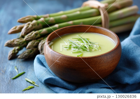 Delicious creamy asparagus soup with melted cheese on blue cloth napkin on wooden table background. Bright green bowl of asparagus andellek soup made with cream or coconut milk with crackers, sprouts 130026836