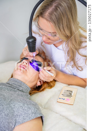 Eyelash extension procedure using ultraviolet lamp in modern beauty salon. Innovative lash enhancement technology with precise application technique. Eyelash extension procedure using ultraviolet lamp in modern beauty salon. Innovative lash enhancement technology with precise application technique. 130027175