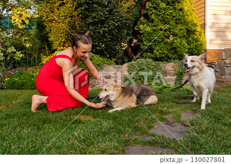 Woman kneeling petting two dogs in lush garden setting Woman kneeling petting two dogs in lush garden setting 130027801