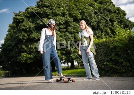 Two friends enjoying a sunny day while trying out a skateboard together Two friends enjoying a sunny day while trying out a skateboard together 130028116