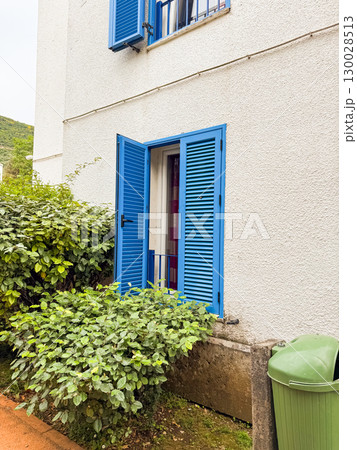 Open blue shuttered window on white building with surrounding bushes and greenery. Residential architecture, lifestyle, eco balance and summer outdoor neighborhood environment. 130028513