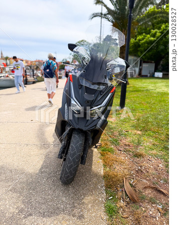 Black scooter with windshield parked on a coastal promenade near people and palm. Urban lifestyle, freedom, holiday transport and modern travel mobility. 130028527