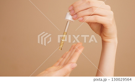Close-up of female hands on isolated beige background holding dropper, anti-aging facial serum, booster dripping on palm.Commercial skincare, moisturizing, vitamin c, anti-wrinkle products advertising 130029564
