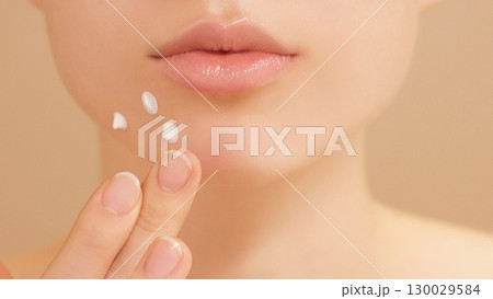 Close-up of a young woman gently applying acne spot cream for clear radiant skin. Self care, skincare, and self-confidence with targeted blemish treatment. Teenage girl, beauty tips on light backdrop 130029584