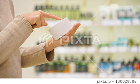 Woman holding a cosmetic jar in a beauty store, reading the label and examining ingredients. Moisturizing anti-aging face cream or nourishing hair mask. Concept of conscious choice, skincare, haircare 130029588