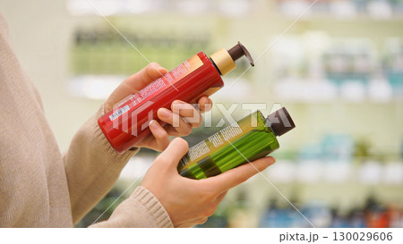 Customer comparing haircare products in a beauty store. Woman reading ingredient labels on bottles of shampoo, conditioner or balm. Concept of conscious choice and personal care for hair and scalp. 130029606