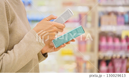 Woman selecting a cosmetic serum for face or hair in a beauty store, reading ingredients, comparing products. Female customer holding skincare or haircare item. Concept of conscious choice, self-care. 130029613