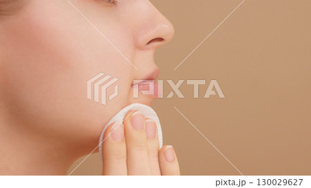 Close-up half face portrait of smiling young woman using cotton pad in studio on beige backdrop. Adorable Caucasian girl using cotton round cleansing skin. Concept of skin care advertising. Copy space Close-up half face portrait of smiling young woman using cotton pad in studio on beige backdrop. Adorable Caucasian girl using cotton round cleansing skin. Concept of skin care advertising. Copy space 130029627