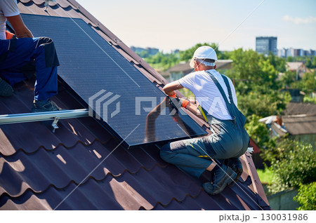Workers building solar panel system on rooftop of house. Two men installers in helmets installing photovoltaic solar module outdoors. Alternative, green and renewable energy generation concept. Workers building solar panel system on rooftop of house. Two men installers in helmets installing photovoltaic solar module outdoors. Alternative, green and renewable energy generation concept. 130031986